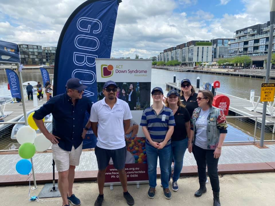 Group of people standing in front of a GoBoat and Down Syndrome Association sign on a wharf 